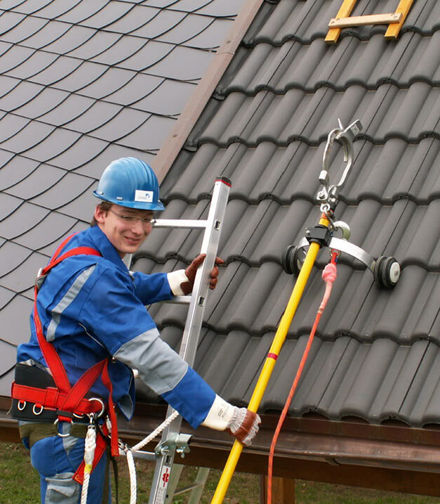 Ein bauarbeiter in Sicherheitsausrüstung steht links auf einer Leiter, die an ein Hausdach gelehnt ist. Er hält in der rechten Hand eine Sicherungsstange.