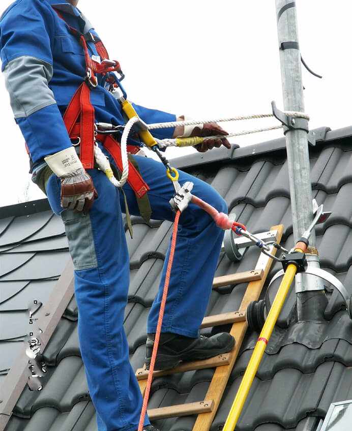 Ein Arbeiter in blauer Kleidung steht auf einem dunklen Hausdach und hält mit einer Hand ein Sicherungsseil, das an seiner Schutzausrüstung befestigt ist.