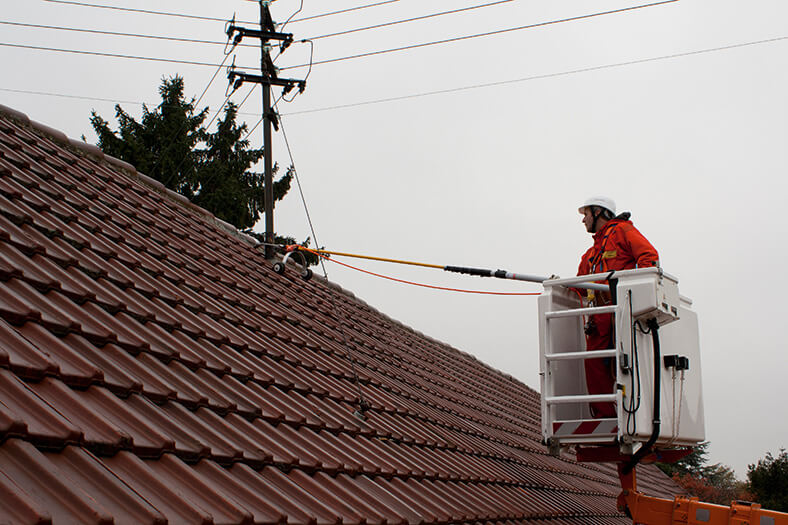 Das Bild zeigt ein Hausdach mit roten Ziegeln, auf dem First eine Antenne, an der Dachschräge sieht man eine Sicherungsstange, rechst ein Bauarbeiter mit Helm in einem Korb.