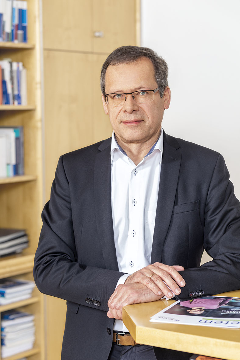 Porträtfoto von Johannes Tichi, Vorsitzender der Geschäftsführung der BG ETEM. Er hat kurze Haare und eine Brille. Er trägt ein dunkles Jackett, ein weißes Hemd und stützt einen Arm auf einen Stehtisch, auf der die neue Ausgabe des etem-Magazins liegt.
