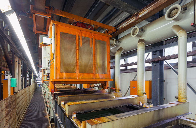 Das Bild zeigt eine automatische Gestellanlage für Galvanotechnik. Man sieht große, weiße Rohre und orangefarbene hängende Elemente entlang einer Fertigungsstraße in einer Werkshalle.