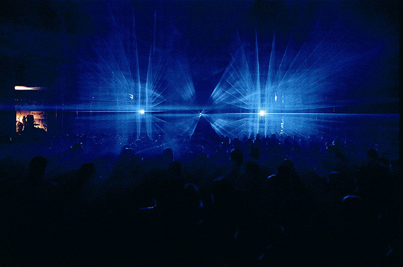 Blaue Laserstrahlen erleuchten von der Bühne aus einen dunklen Saal, in der unteren Hälfte des Bildes sieht man dunkle Umrisse von Personen in einer Menschenmenge. 