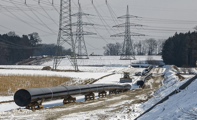 Baus eines großen, oberirdisch verlaufenden Gasleitungsrohres in winterlicher Landschaft, im Hintergrund Strommasten mit Überland-Leitungen.