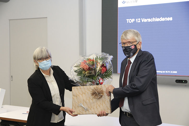 Das Foto zeigt links Karin Jung und rechts Dr. Heinz-Willi Mölders. Sie halen zwischen sich einen Blumenstrauß und ein verpacktes Geschenk. Frau Jung hat kürzere graue Haare, trägt eine Brille und einen blauen Mund-Nasen-Schutz, eine helle Bluse und einen schwarzen Blazer, Dr. Mölders hat graue Haare, trägt eine Brille sowie einen schwarz-grauen Mund-Nasen-Schutz, einen grauen Anzug mit roter Krawatte und hellem Hemd.