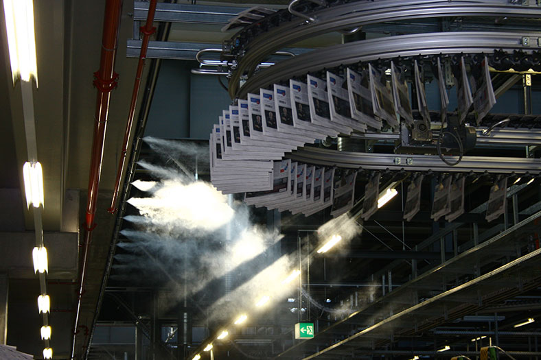 Aus einer Rohrleitung links strömt aus mehreren Öffnungen Wasserdampf in eine Produktionshalle. An einem Karussell rechts hängen Druckerzeugnisse.