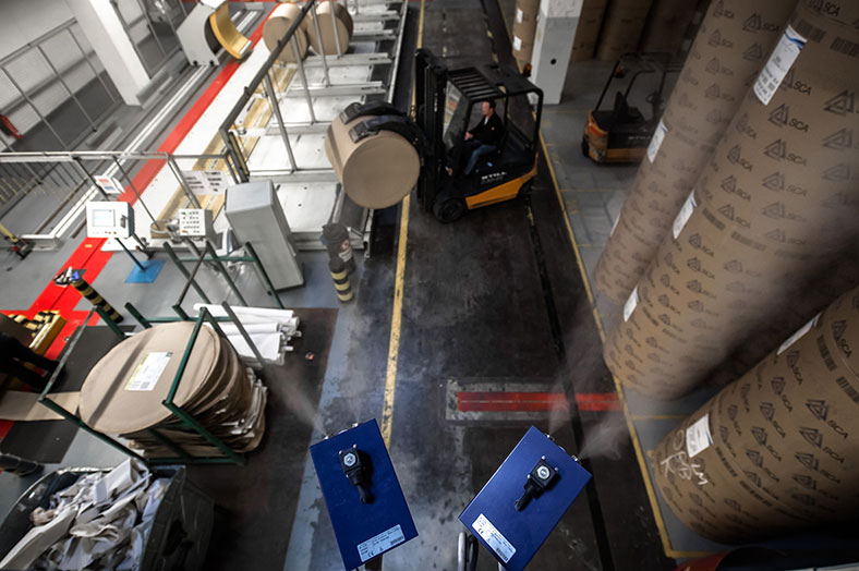 Zwei blaue Luftbefeuchter im Vordergrund verströmen von der Decke aus Wasserdampf in eine Werkshalle. Im Bild rechts stehen Stapel von Zylindern mit Papier, in der MItte transportier ein Gabelstapler einen Zylinder.