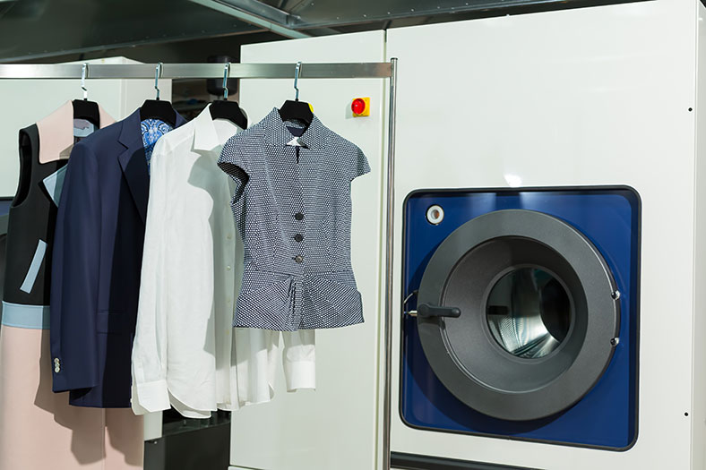 Rechts eine industrielle Waschmaschine, rechts eine Kleiderstange, an der verschiedene Kleidungsstücke an Kleiderbügeln hängen.