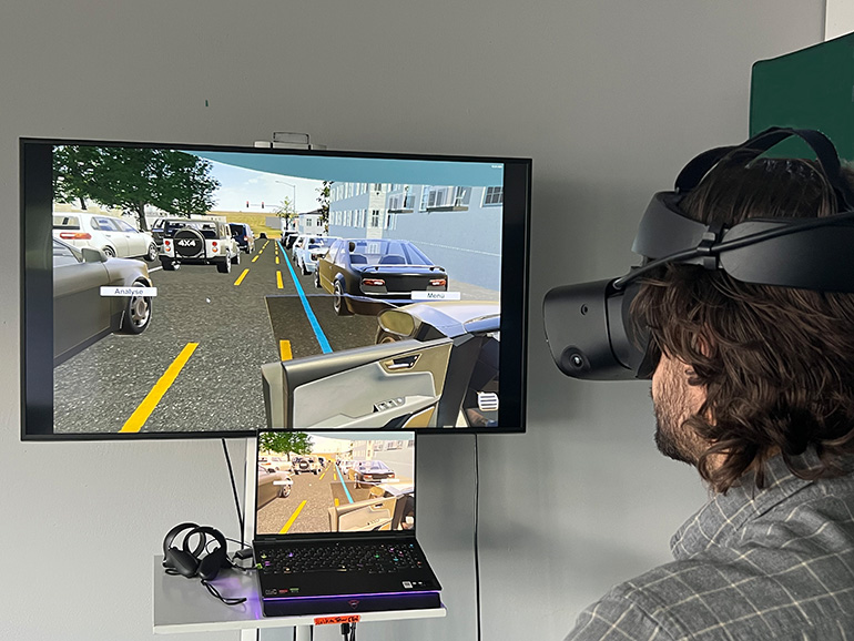 Mann mit dunklen Haaren trägt eine VR-Brille und schaut auf einen größeren Bildschirm mit einer Verkehrssimulation. Darunter liegt auf einem Pult ein Laptop mit einem kleineren Bildschirm, der dieselbe Simulation zeigt.