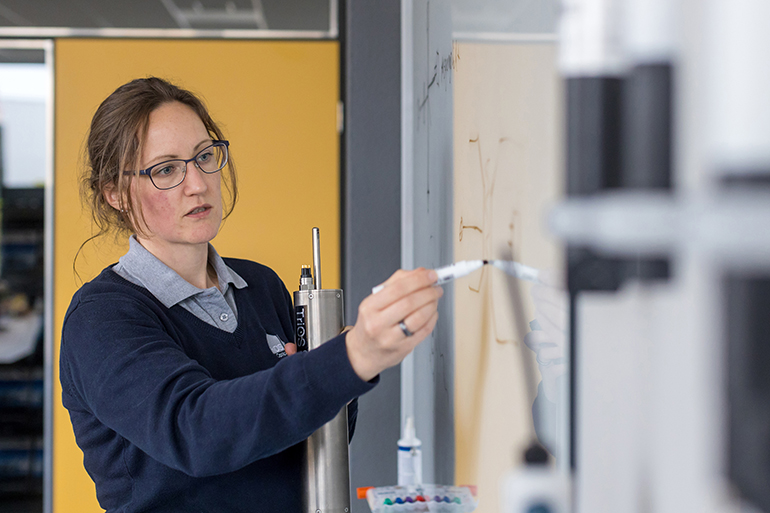 Dr. Alexandra Ostmann steht an einem Whiteboard und zeichnet etwas darauf, in der anderen Hand hält sie ein Messgerät aus Metall. Sie hat zurückgebundene braune Haare, trägt eine Brille und einen dunkelblauen Pullover.