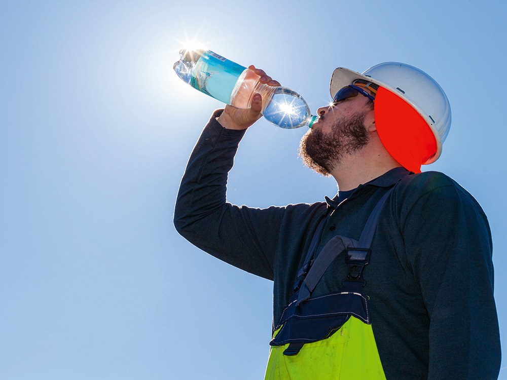 Arbeiter im Freien in Sonnenschutzkleidung mit Helm und Sonnenbrille trinkt aus einer Wasserflasche.