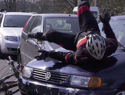 Ein Radfahrer in Stuntman-Montur rollt sich nach einer Kollision mit einem Pkw über die Motorhaube ab; im Hintergrund weitere Fahrzeuge
