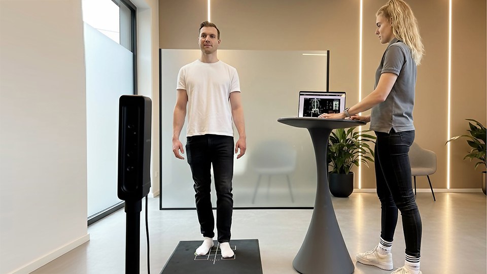 Mann steht auf einer Matte mit Positionsmarkierung, eine spezielle Kamera erfasst seine Körpersilhouette; neben ihm eine Frau an einem Stehtisch, die einen Laptop bedient.