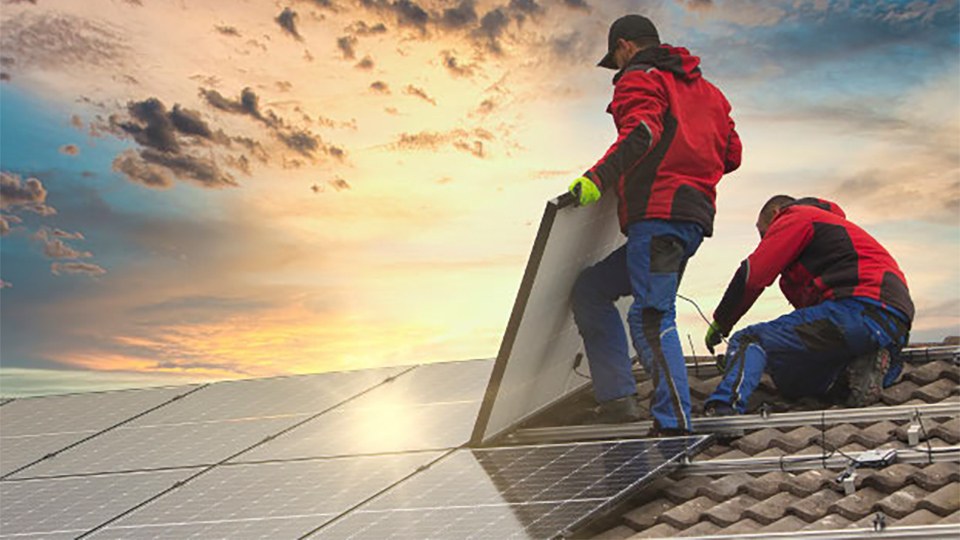 Zwei Arbeiter in Arbeitskleidung bringen auf einem Dach Solarpaneele an.