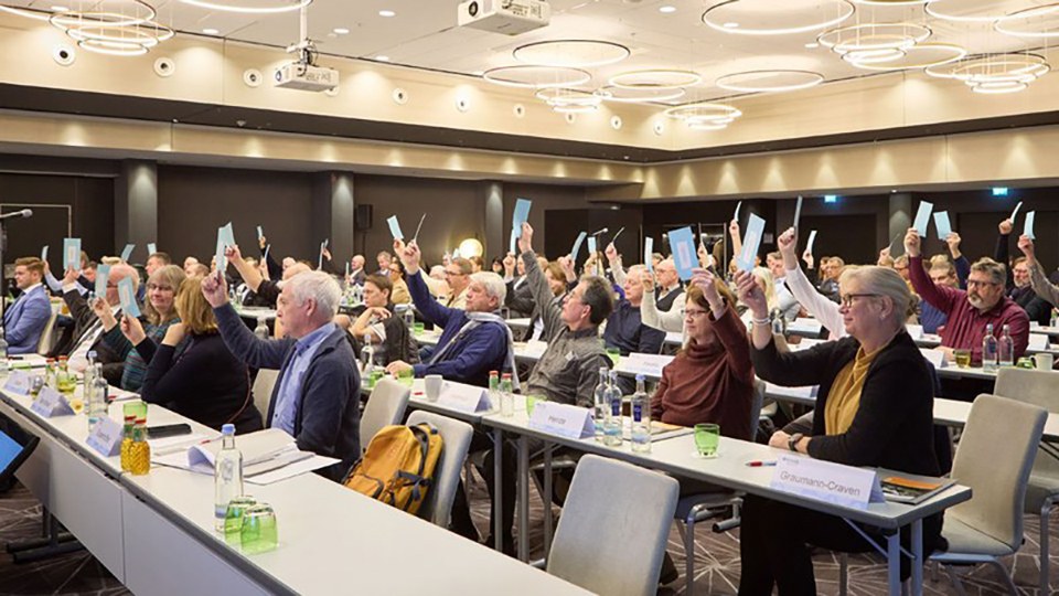 Seitlicher Blick in den Sitzungssaal mit der Vertreterversammlung, die mittels Heben eines Zettels eine Abstimmung durchführt