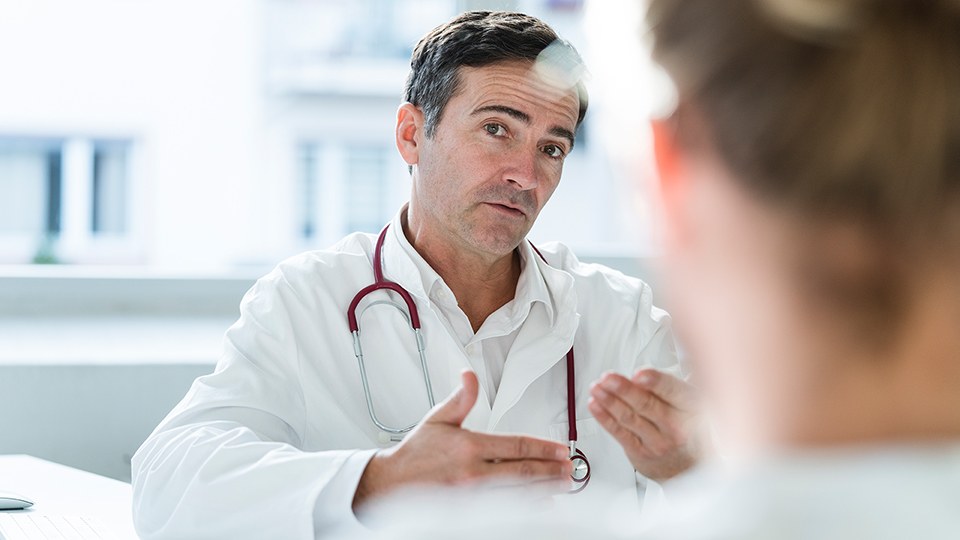 Arzt im weißen Kittel und mit Stethoskop um den Hals sitzt am Schreibtisch im Gespräch mit einer Patientin, die nur unscharf von hinten zu sehen ist.
