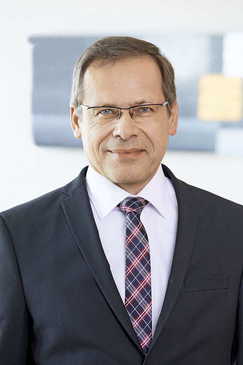 Das Porträtfoto zeigt Johannes Tichi, Vorsitzender der Geschäftsführung der BG ETEM. Er hat kurze Haare und trägt eine Brille. Er trägt ein dunkles Jackett, ein weißes Hemd und eine blau gestreifte Krawatte.