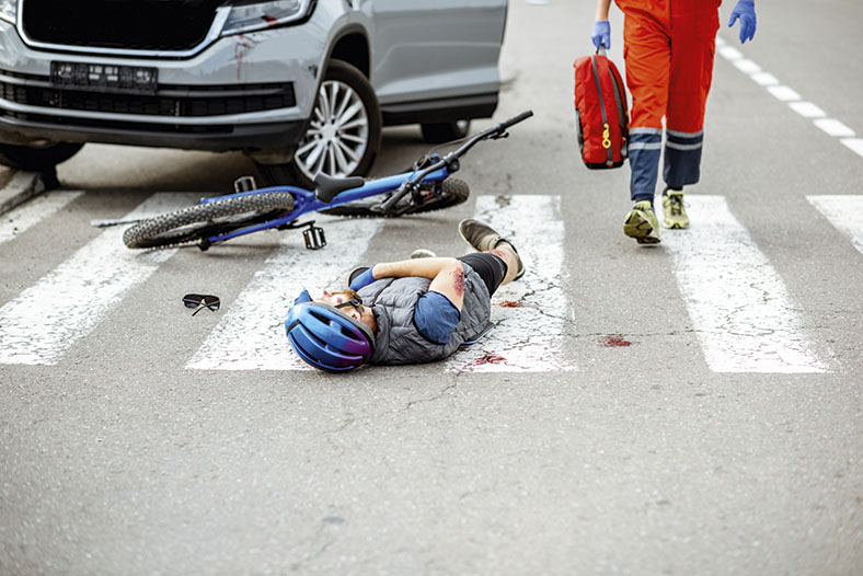 Das Foto zeigt einen verletzten Radfahrer mit Schutzhelm, der auf einem Zebrastreifen liegt. Er blutet am Arm. Blutflecke sind ebenfalls auf dem Zebrastreifen. Vor ihm das Vorderrad seines Fahrrads.