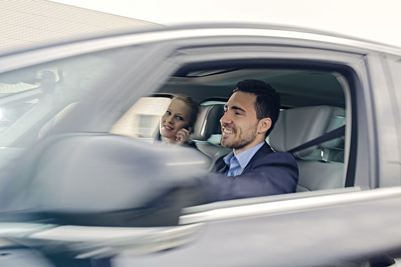 Ein Mann sitzt bei geöffnetem Fenster in einem Pkw. Eine Frau sitzt auf dem Beifahrersitz und lächelt.
