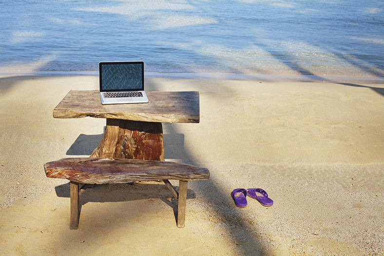 Dieses Foto zeigt einen Holztisch auf dem ein Laptop steht. Davor steht eine Holzbank und daneben ein paar lila Badeschuhe. Im Hintergrund erkennt man das Meer.