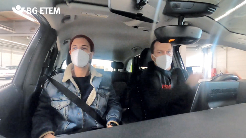 Das Bild zeigt den Innenraum eines Pkw mit zwei Personen, die beide einen weißen Mundschutz tragen. Der Fahrer testet seine Reaktionen unter Alkoholeinfluss.