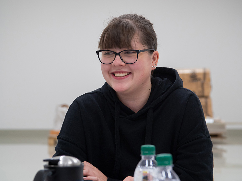 Das Bild zeigt das Porträt einer lächelnden Jugendlichen mit Brille. Ihre dunklen Haare sind zurückgebunden. Sie sitzt an einem Tisch, vor ihr sind Getränkeflaschen und eine Kaffeekanne teilweise sichtbar.