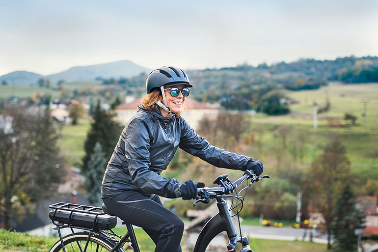 Schutzhelme für E-Biker: Frau in schwarzer Kleidung, mit Sonnenbrille und schwarzem Helm fährt auf einem E-Bike durch die Natur.