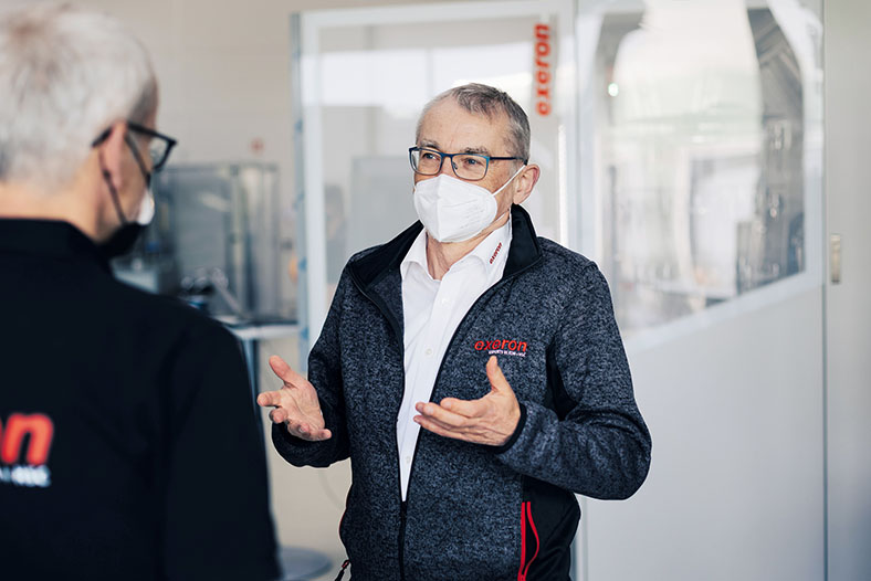 Post-Covid: Unternehmer Bernd Fleiner nach seiner Covid-Erkrankung im Gespräch mit einem Mann mit grauen Haaren und Brille in schwarzer Kleidung und schwarzer FFP2-Maske. Herr Fleiner hat kurze graue Haare, trägt einen Bart, eine Brille, eine weiße FFP2.Maske ein weißes Hemd und eine grau-schwarze Strickjacke.
