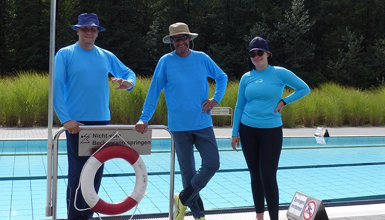 3 Personen stehen vor einem Freibad-Becken. Sie tragen hellblaue Langarm-Shirts und lange Hosen, Sonnenhüte und Sonnenbrillen.