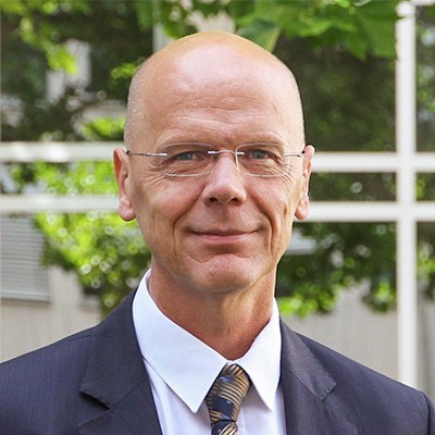 Portrait von Dr. Johannes Hüdepohl, BG ETEM. Herr Hüdepohl steht im Freien, er hat eine Glatze, trägt eine Brille und einen dunklen Anzug mit weißem Hemd und Krawatte. 