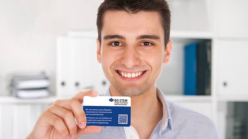 Ein junger Mann präsentiert lächelnd eine Versichertenkarte der BG ETEM.
