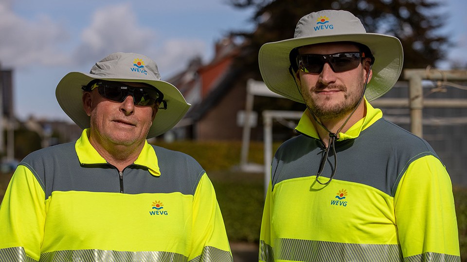 Zwei Beschäftigte von WEVG; sie tragen Sonnenbrillen sowie Sonnenschutzhüte und langärmelige Oberteile in Signalfarbe, auf denen Logo und Schriftzug des Unternehmens aufgebracht sind; unscharf im Hintergrund Häuser eines Wohngebiets.