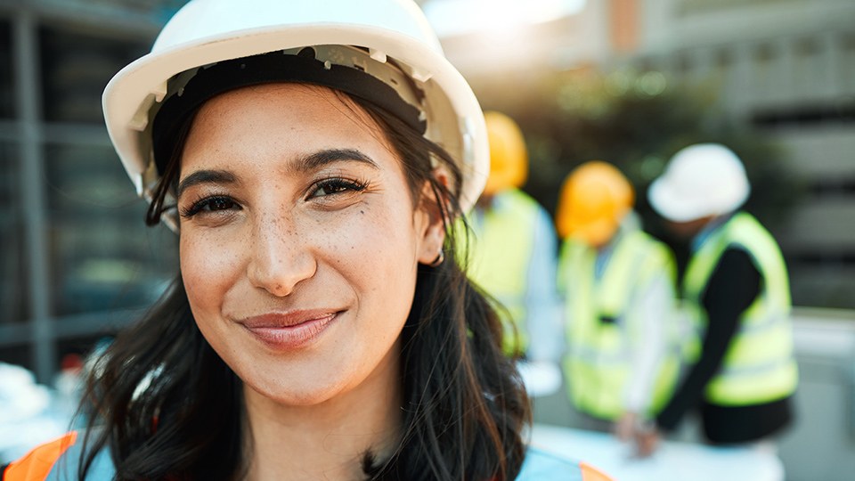 Junge Frau mit Schutzhelm und Sicherheitsweste auf einer Baustelle, sie lächelt in die Kamera, hinter ihr sind unscharf drei Kollegen zu erkennen