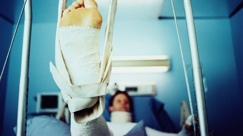 Mann liegt in einem Krankenhausbett, er trägt eine Halskrause und ist nur unscharf zu erkennen, im Vordergrund des Bildes ist groß sein bandagierter Fuß, zu sehen, der in einer Beinschlinge hängt.
