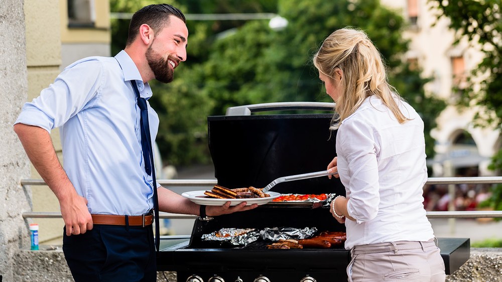 Ein Mann und eine Frau in lockerem Büro-Outfit an einem Gasgrill im Außenbereich eines Gebäudes; die Krawatte des Mannes ist gelockert; er hält eine Platte mit Grillgut und lächelt die Frau an, die weiteres Grillgut auf die Platte legt.