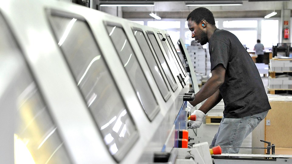 Ein Arbeiter steht an in einer Druckerei an einer eingehausten Weiterverarbeitungsanlage.