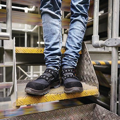 Arbeiter, der nur bis zu den Knien im Bild ist, in Jeans und mit Sicherheitsschuhen auf einer Treppe in einer Fabrikhalle.