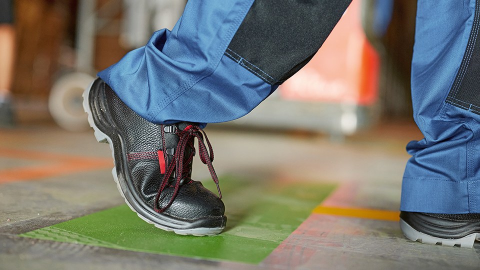 Ein Arbeiter, von dem nur die Sicherheitsschuhe und die Sicherheitshose bis zu den Knien zu sehen sind, geht über den Boden einer Fabrik