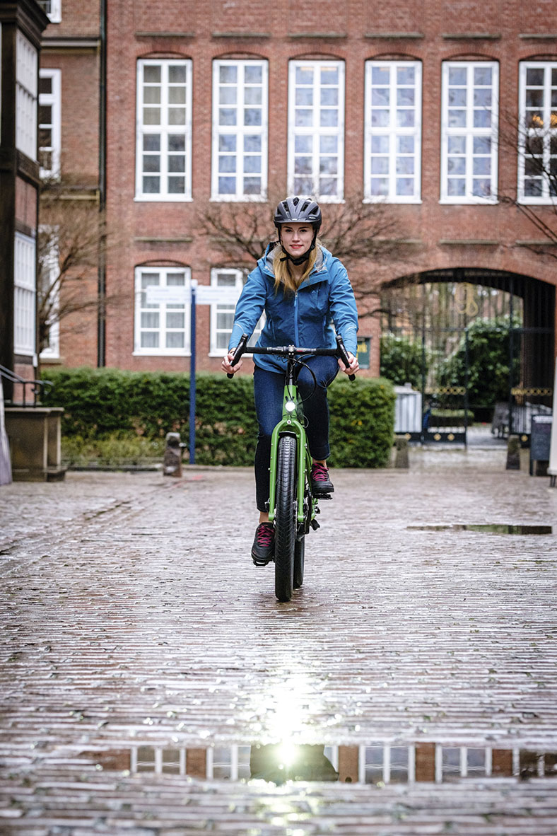Frontalansicht einer Radfahrerin mit dunklem Schutzhelm und blauer Jacke auf einem grünen Fahrrad. Im Hintergrund ein roter Backsteinziegelbau mit weißen Sprossenfenstern.