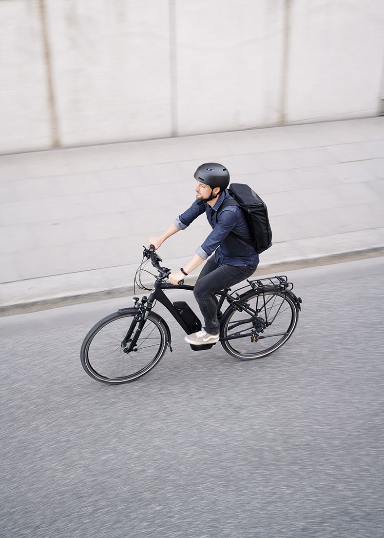 Ein Radfahrer in Jeanshose und -hemd mit schwarzem Fahrradhelm und Rucksack fährt auf einem schwarzen Fahrrad eine leere Straße entlang, Ansicht von schräg oben.