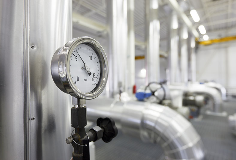 Gasdruckregler mit runder Gasmanometeranzeige in einem Gaswerk, im Hintergrund Gasleitungen mit Thermoschutzummantelung.