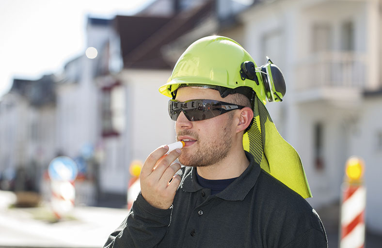 Arbeiter mit Schutzhelm und Nackenschutz steht draußen im Sonnenschein, er trägt eine Sonnenbrille und trägt mit einem Lippenstift UV-Schutzmittel auf.