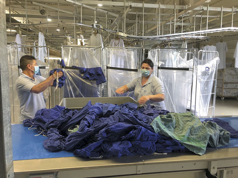 Eine Mitarbeiterin und ein Mitarbeiter der Firma Bardusch Textil Mietdienste GmbH in Augsburg sortieren dunkelblaue Textilien auf einem Tisch. Beide tragen einen Mund-Nasen-Schutz.
