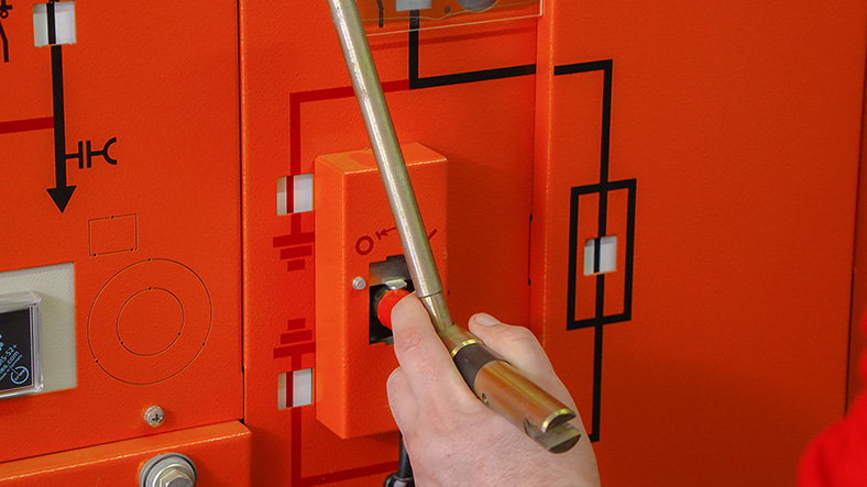 Auf dem Bild ist eine elektrische Schaltanlage in orange zu sehen. Eine Hand dreht mit einem Stiftschlüssel an einem Entriegelungskästchen.