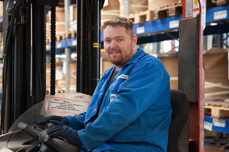 Maschinen- und Anlagenführer Christian Marcus steht in blauer Arbeitskleidung in einer Lagerhalle mit Drahtspulen.