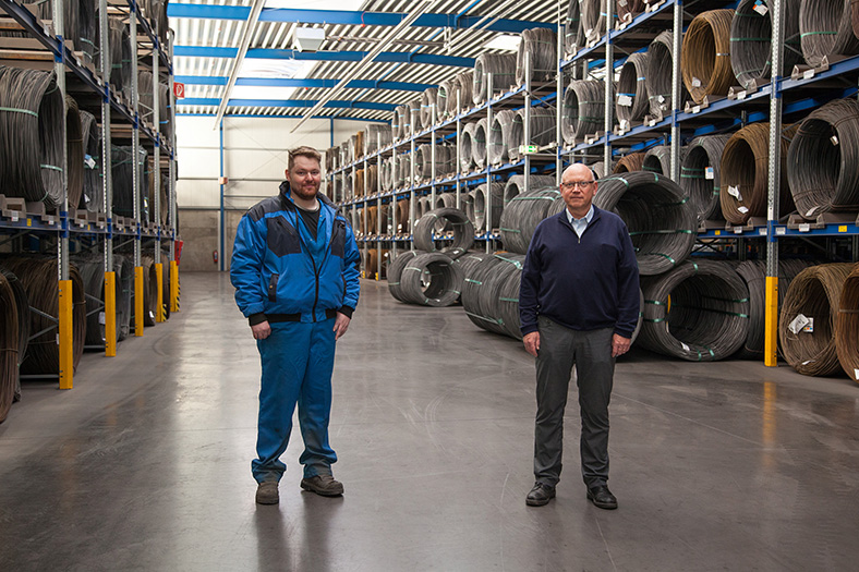 Maschinen- und Anlagenführer Christian Marcus und technischer Leiter Albrecht Borner stehen nebeneinander in einer Lagerhalle mit Regalen voller Drahtspulen.