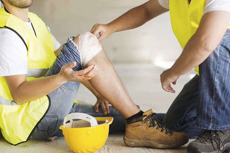 Ein Bauarbeiter mit gelber Warnweste und einer einbandagierten Wunde am Knie sitzt auf dem Boden. Ein weiterer Bauarbeiter kniet vor ihm und versorgt die Wunde.