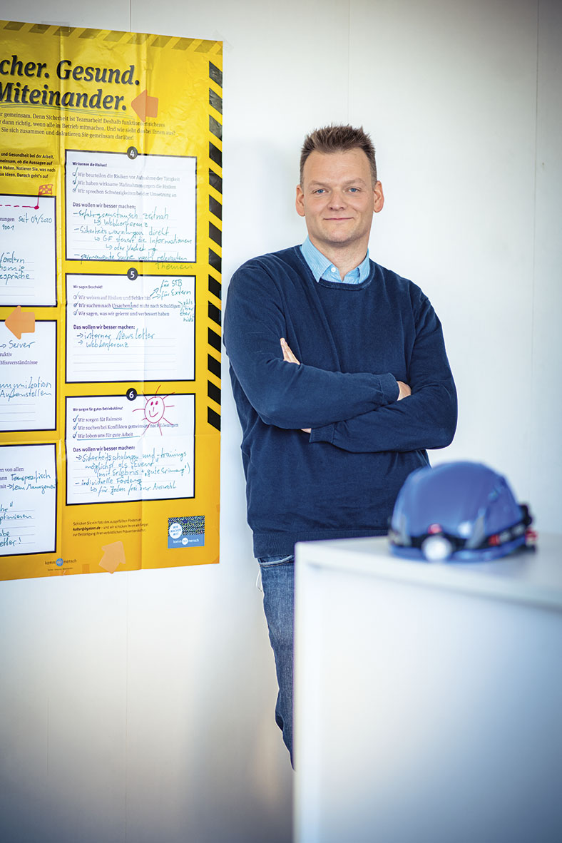 Elektroingenieur und Sicherheitsfachkraft Raiko Schmidt von der STB Sachsenwind steht in einem blauen Pullover mit verschränkten Armen neben dem gelben Risikoposter der BG ETEM.
