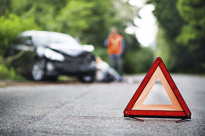 Gesicherte Unfallstelle: Rotes Warndreieck steht im Vordergrund, dahinter ein verbeulter Unfallwagen mit zwei Personen, eine sitzend, eine stehend.