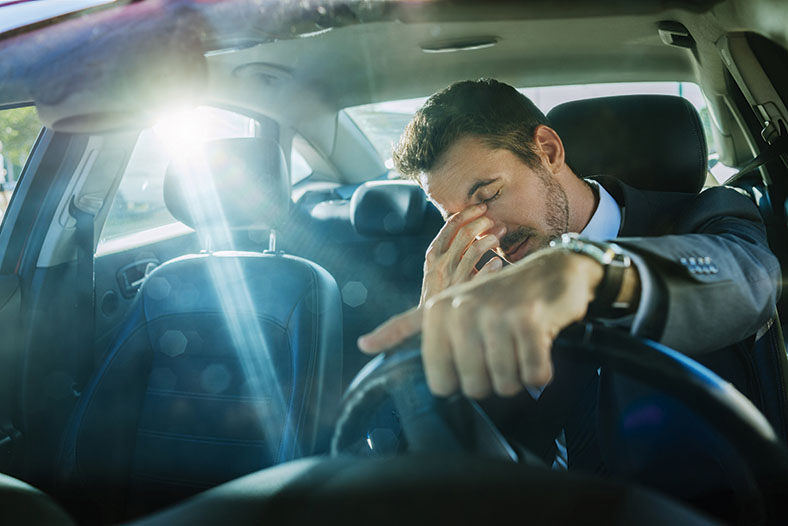 Ermüdeter Autofahrer sitzt am Steuer, hat die Augen geschlossen und greift sich mit den Fingern an die Nasenwurzel. 