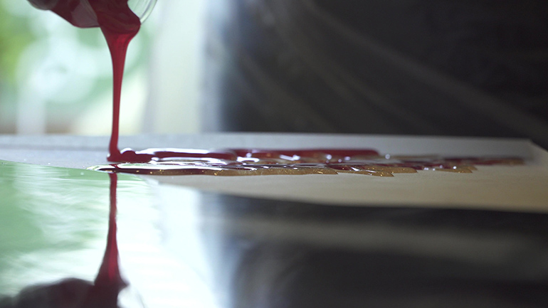 Eine dickflüssige rote Substanz wird aus einem Glas auf einen glatten, glänzenden Untergrund geschüttet.